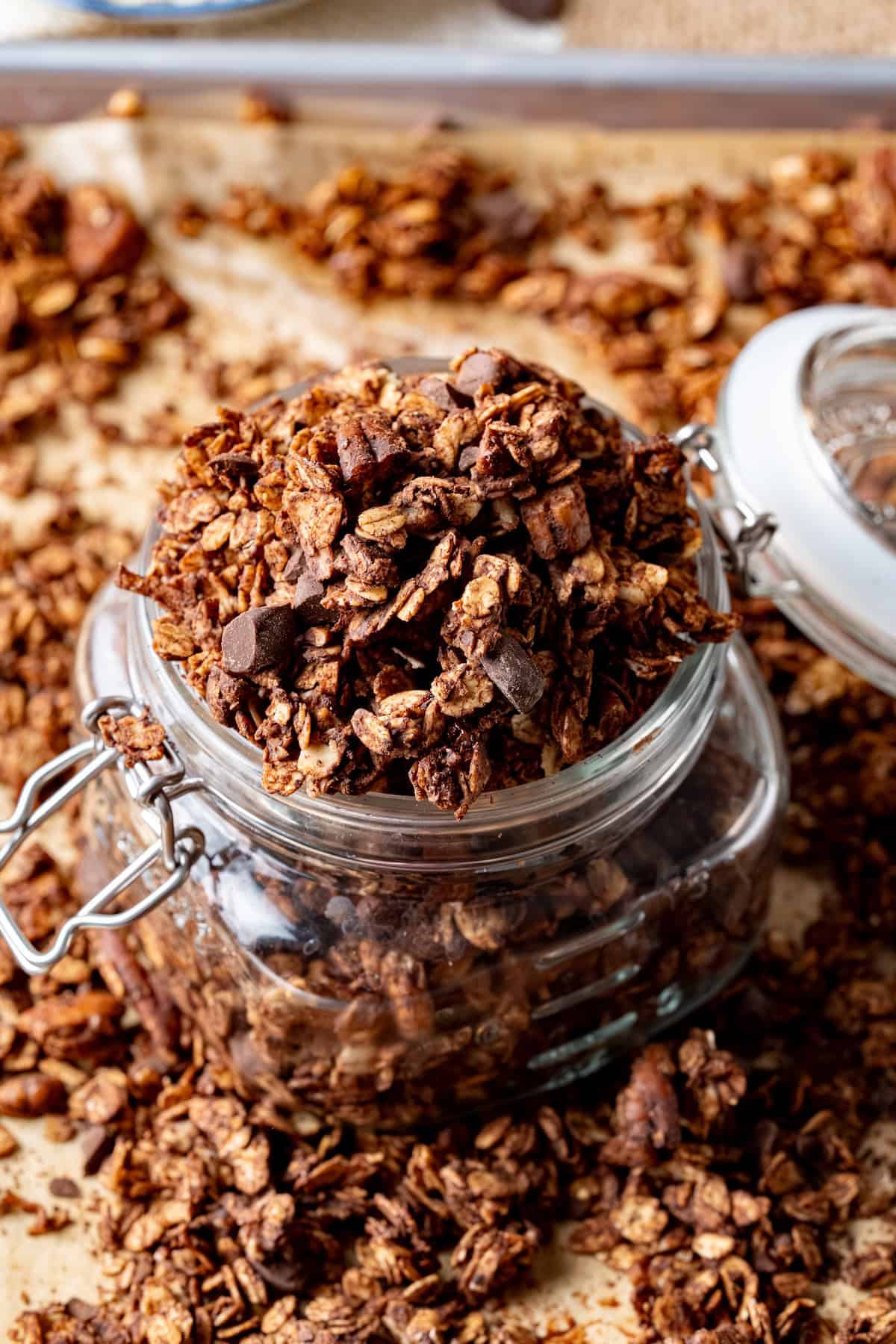 Glass jar filled with homemade chocolate coconut granola made from oats, pecans, and dark chocolate chunks, with extra granola scattered on parchment paper in the background.