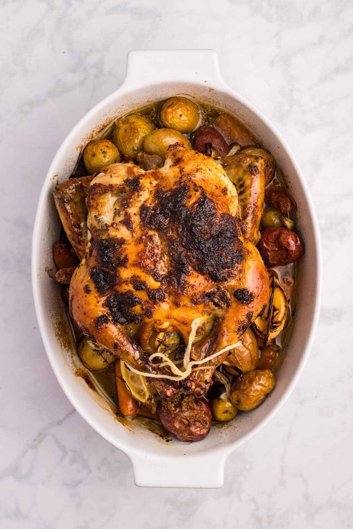 Golden brown herb roasted chicken resting on a bed of roasted baby potatoes, carrots, and lemon wedges in a white baking dish, fresh from the oven.