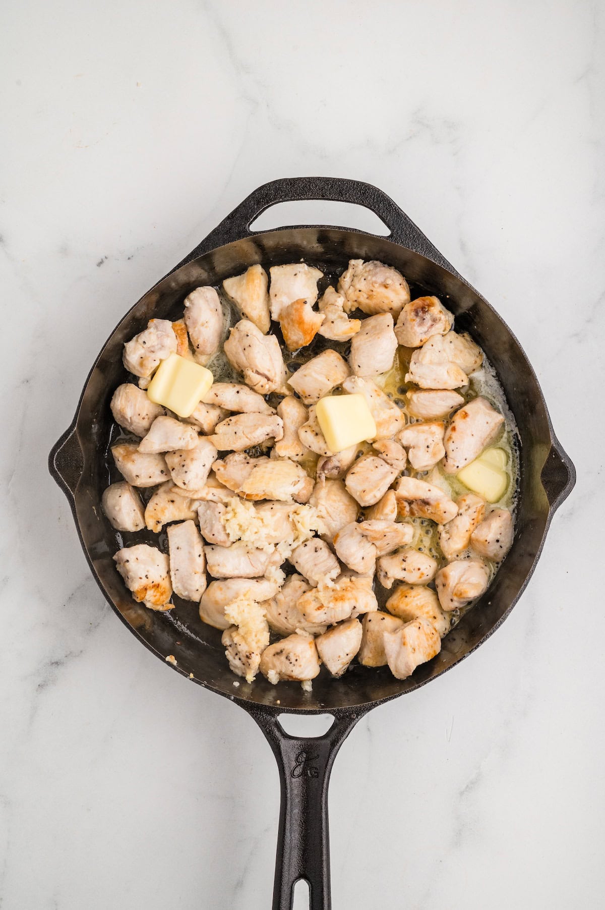Top-down view of browned chicken pieces cooking in a cast iron skillet with melted butter and freshly minced garlic. The chicken is golden on the edges, sizzling in a buttery garlic sauce, as part of the process for making One-Pan Garlic Parmesan Chicken Bites.