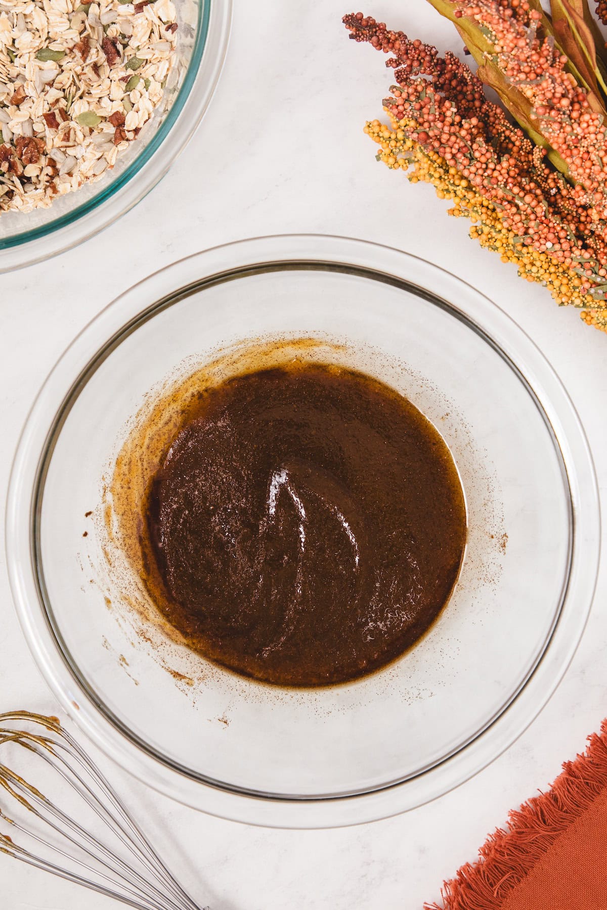 Glass bowl showing blended pumpkin puree mixture with maple syrup, olive oil, and spices forming a smooth, dark pumpkin spice coating, ready to be combined with oats and seeds.