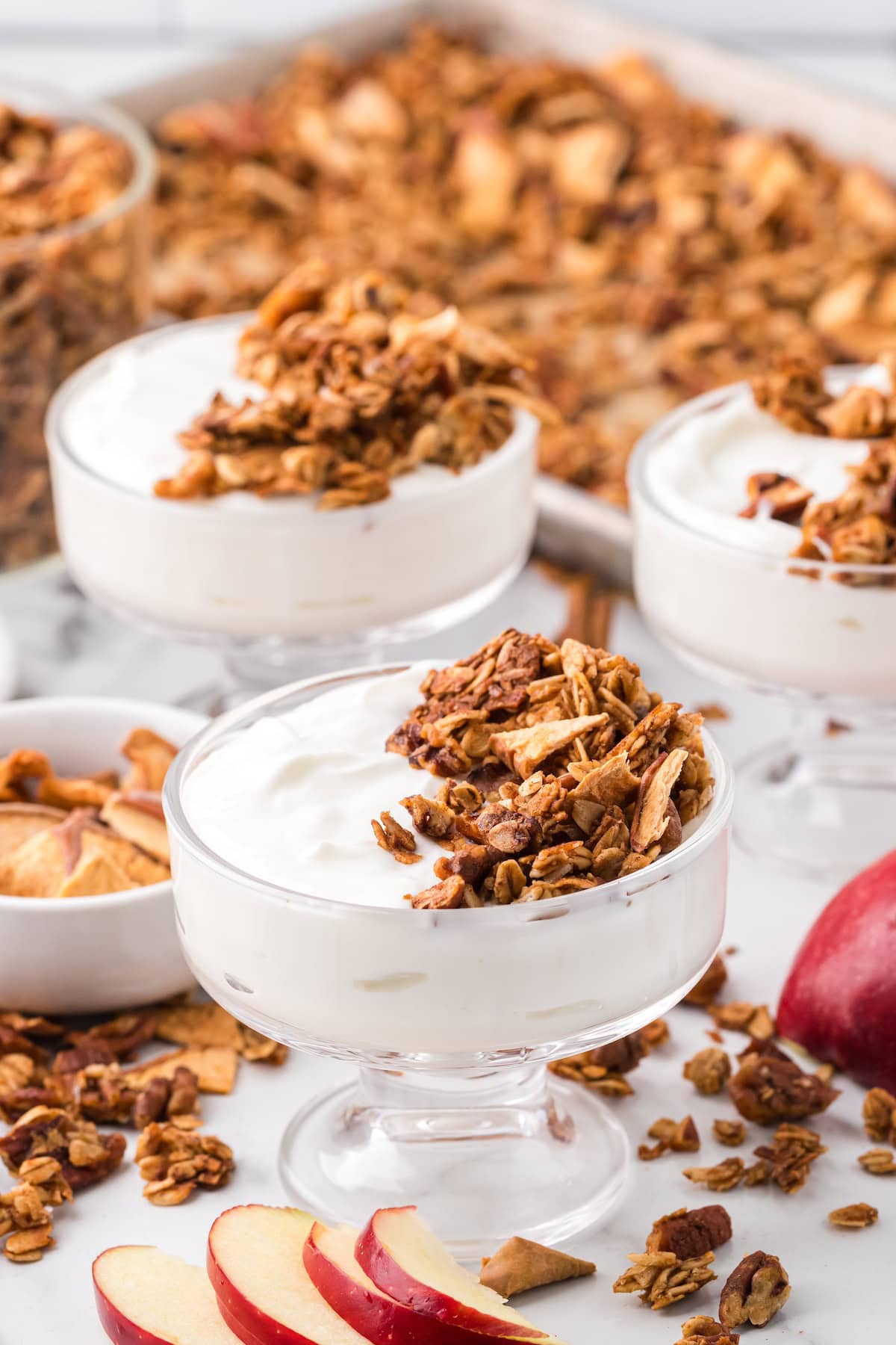 Glass bowls filled with creamy yogurt topped with homemade apple pie granola made with oats, pecans, walnuts, and dried apples, surrounded by sliced fresh apples and a baking tray of granola in the background.