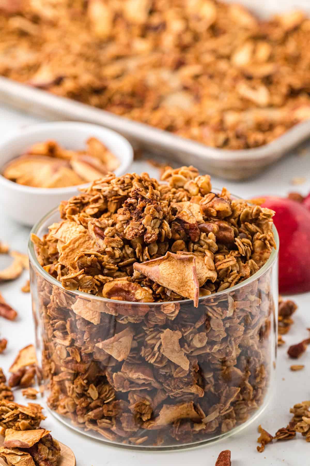 Glass jar filled with crunchy homemade apple pie granola with dried apple slices, walnuts, and pecans, with a baking tray of granola in the background.