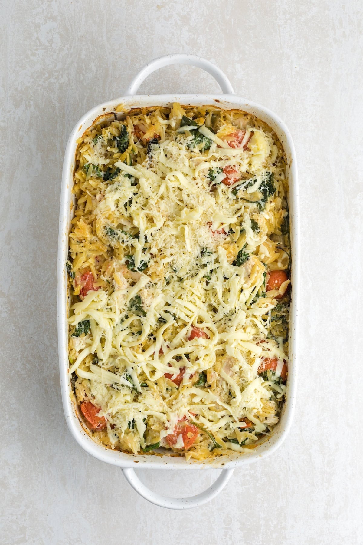 Baked creamy chicken orzo casserole fresh out of the oven, topped with melted cheese, spinach, and cherry tomatoes in a white baking dish.