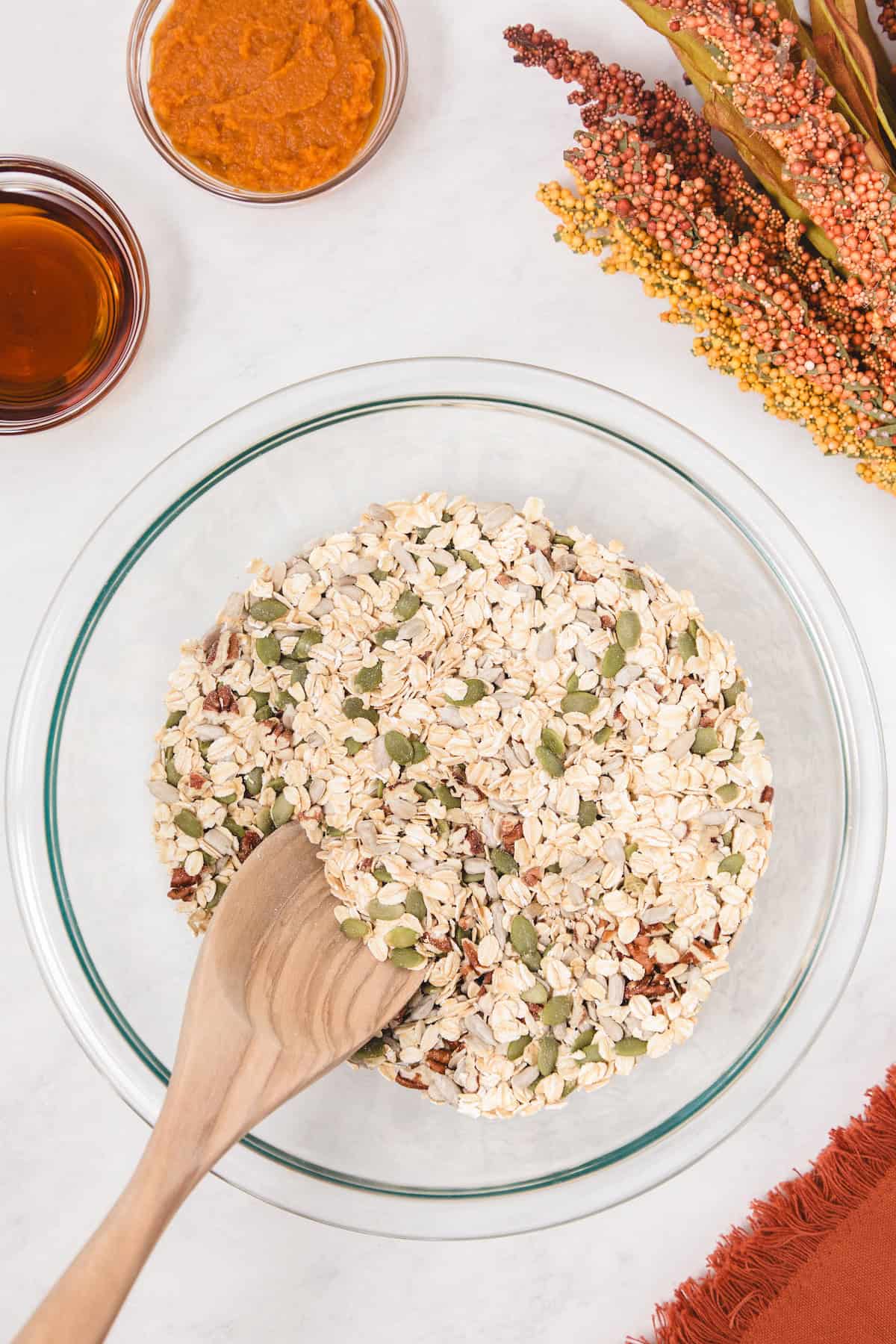 Wooden spoon mixing rolled oats, pumpkin seeds, sunflower seeds, and chopped pecans in a glass bowl, with small bowls of pumpkin puree and maple syrup nearby on a white surface decorated with autumn-colored flowers.