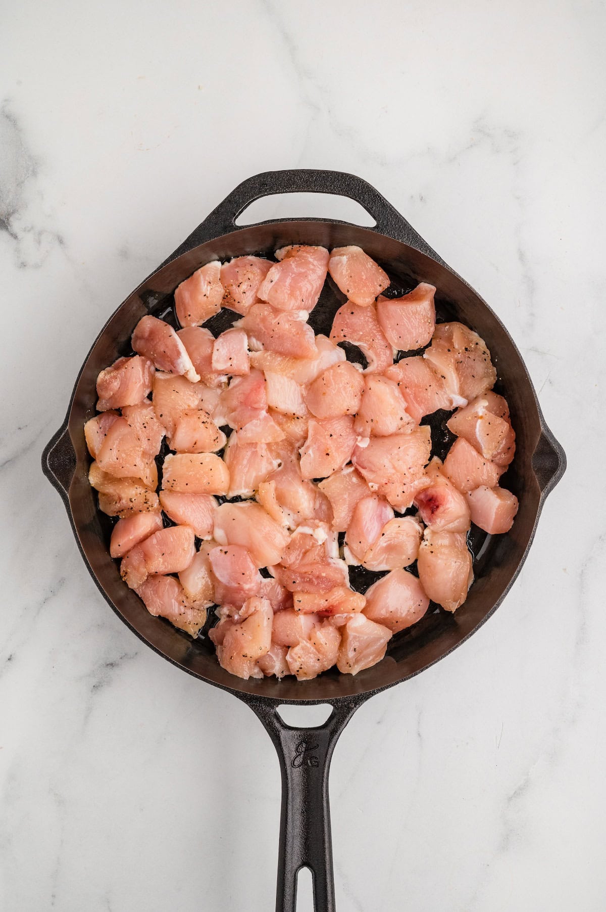 Top-down view of seasoned raw chicken pieces spread evenly in a cast iron skillet with avocado oil, ready to be cooked for One-Pan Garlic Parmesan Chicken Bites on a marble surface.