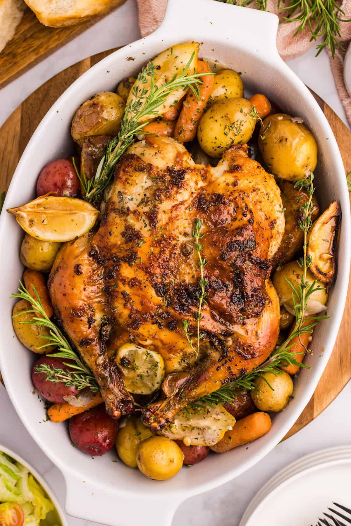 Golden herb roasted chicken surrounded by baby potatoes, carrots, and lemon wedges in a white baking dish, garnished with fresh rosemary and thyme.