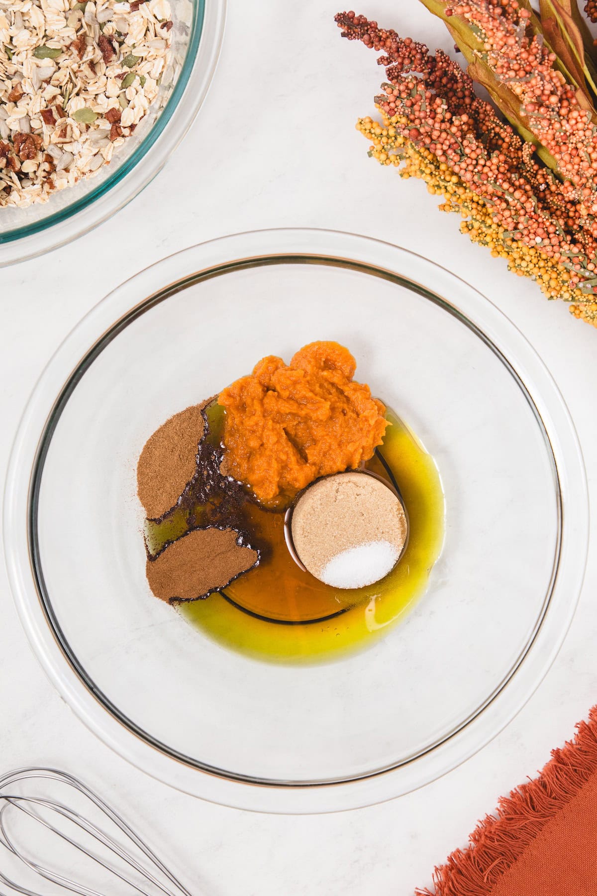 Glass bowl filled with pumpkin puree, maple syrup, olive oil, coconut sugar, pumpkin pie spice, cinnamon, and salt before mixing, with a whisk nearby and a bowl of oats and seeds in the background.