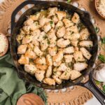 Top-down view of One-Pan Garlic Parmesan Chicken Bites in a cast iron skillet. The golden, tender chicken pieces are coated in a buttery garlic parmesan sauce and garnished with chopped parsley. Surrounded by small bowls of shredded parmesan, fresh salad, a garlic bulb, and a wooden spoon on a green linen napkin for a cozy, rustic presentation.