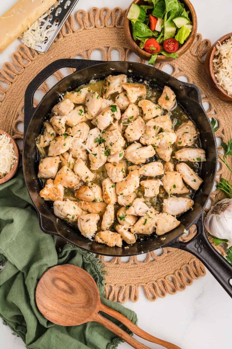 Top-down view of One-Pan Garlic Parmesan Chicken Bites in a cast iron skillet. The golden, tender chicken pieces are coated in a buttery garlic parmesan sauce and garnished with chopped parsley. Surrounded by small bowls of shredded parmesan, fresh salad, a garlic bulb, and a wooden spoon on a green linen napkin for a cozy, rustic presentation.