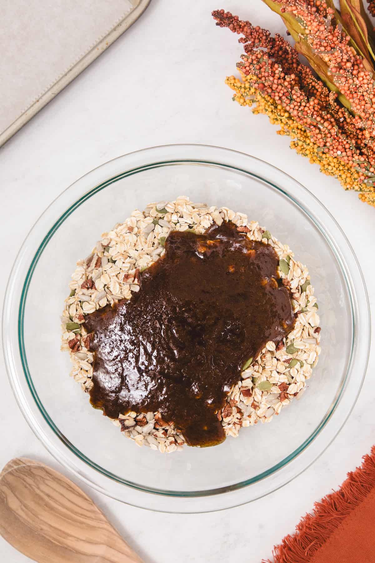 Pumpkin spice mixture made with pumpkin puree, maple syrup, and spices poured over oats, seeds, and chopped pecans in a glass bowl, ready to be mixed together for granola.