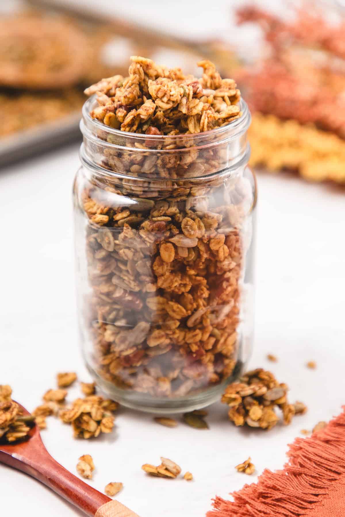 Glass jar filled with crunchy homemade pumpkin spice granola made from oats, nuts, and seeds, with scattered clusters around the jar on a white surface and a warm autumn background