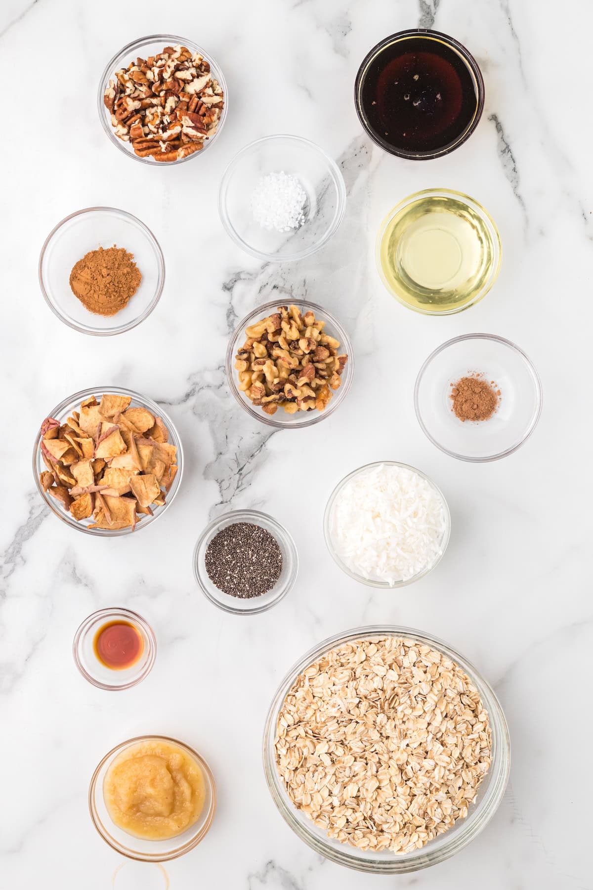 Overhead view of ingredients for homemade apple pie granola including rolled oats, pecans, walnuts, dried apples, coconut flakes, chia seeds, applesauce, maple syrup, coconut oil, cinnamon, nutmeg, and vanilla on a marble surface.