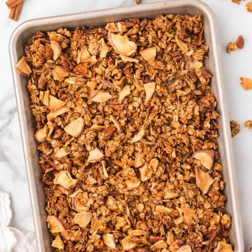 Sheet pan filled with golden homemade apple pie granola made with oats, pecans, and dried apple pieces, cooling on a marble counter with cinnamon sticks beside it.