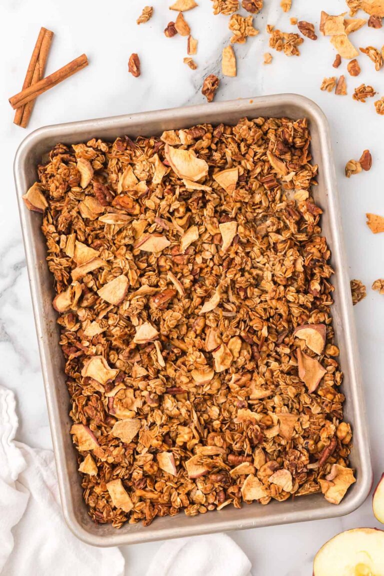 Sheet pan filled with golden homemade apple pie granola made with oats, pecans, and dried apple pieces, cooling on a marble counter with cinnamon sticks beside it.