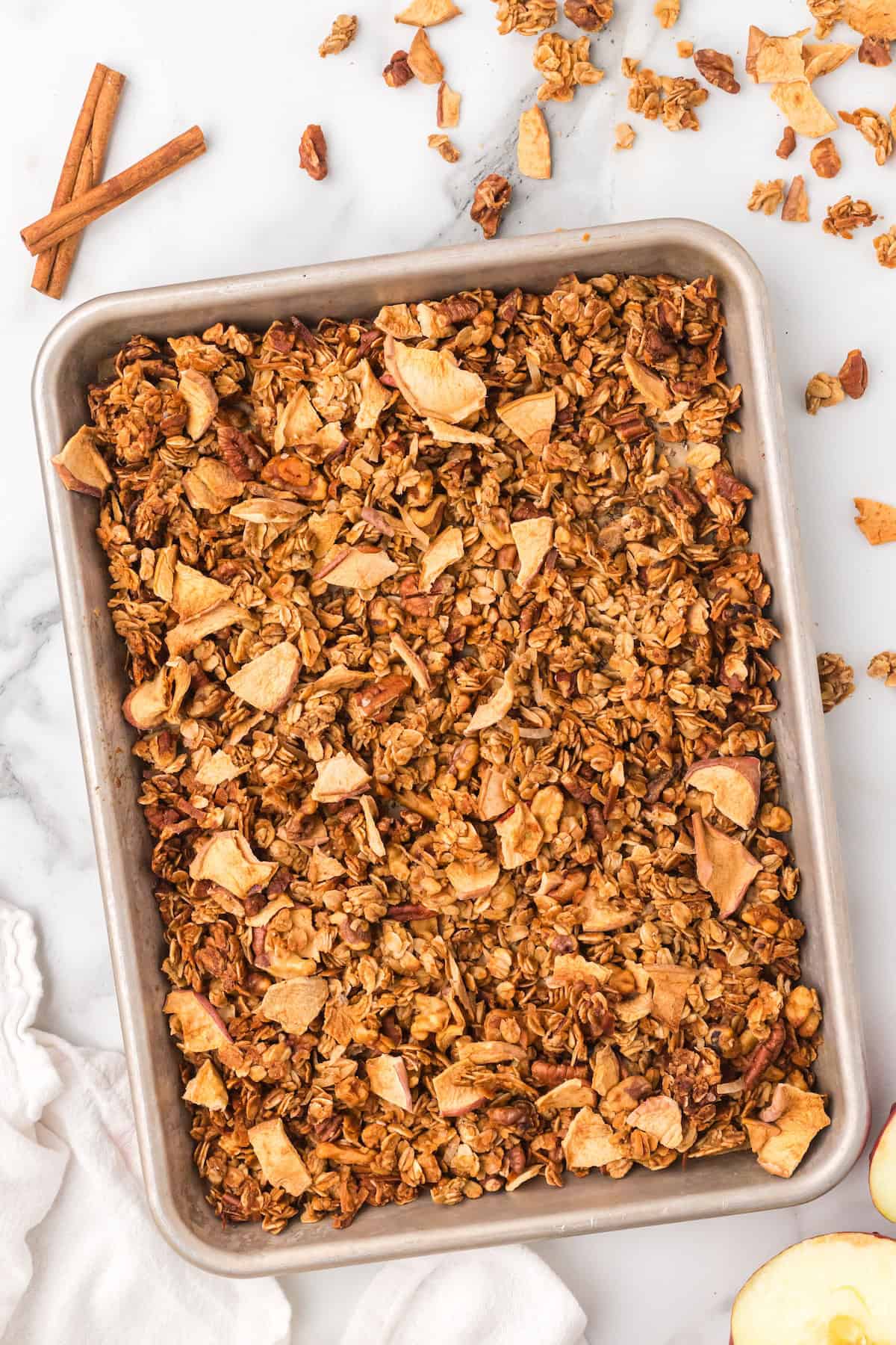 Sheet pan filled with golden homemade apple pie granola made with oats, pecans, walnuts, and dried apple pieces, cooling on a marble counter with cinnamon sticks beside it.