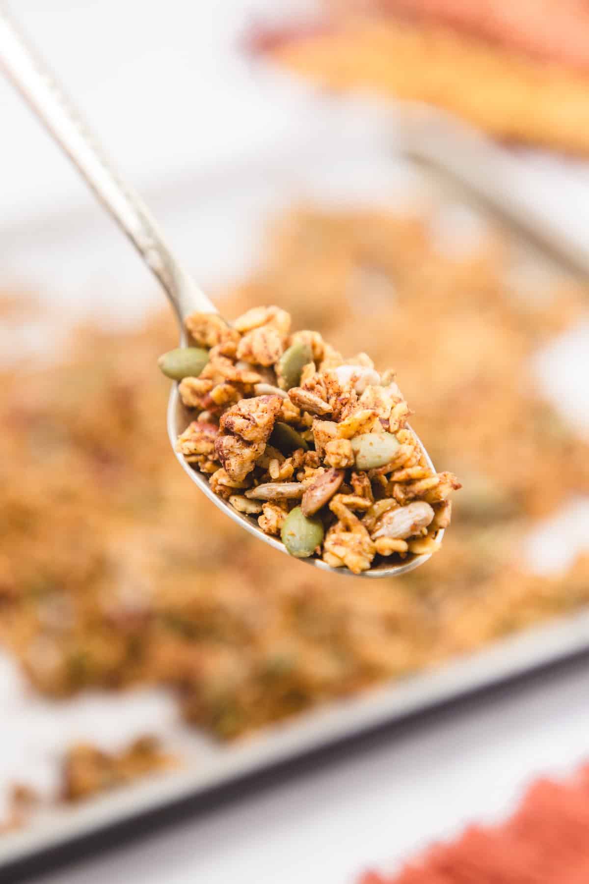 Close-up of a spoonful of homemade pumpkin spice granola showing oats, pumpkin seeds, sunflower seeds, and pecans, with a baking sheet of granola blurred in the background.