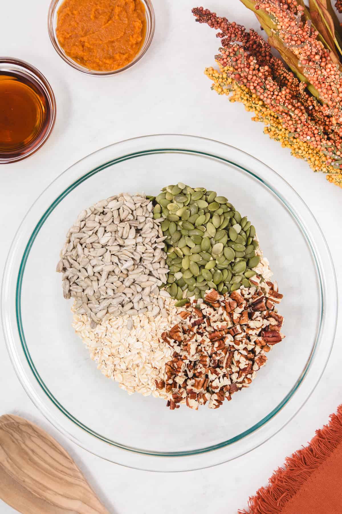 Glass mixing bowl filled with rolled oats, chopped pecans, pumpkin seeds, and sunflower seeds arranged in sections, surrounded by small bowls of pumpkin puree and maple syrup on a white surface with autumn-colored decor.