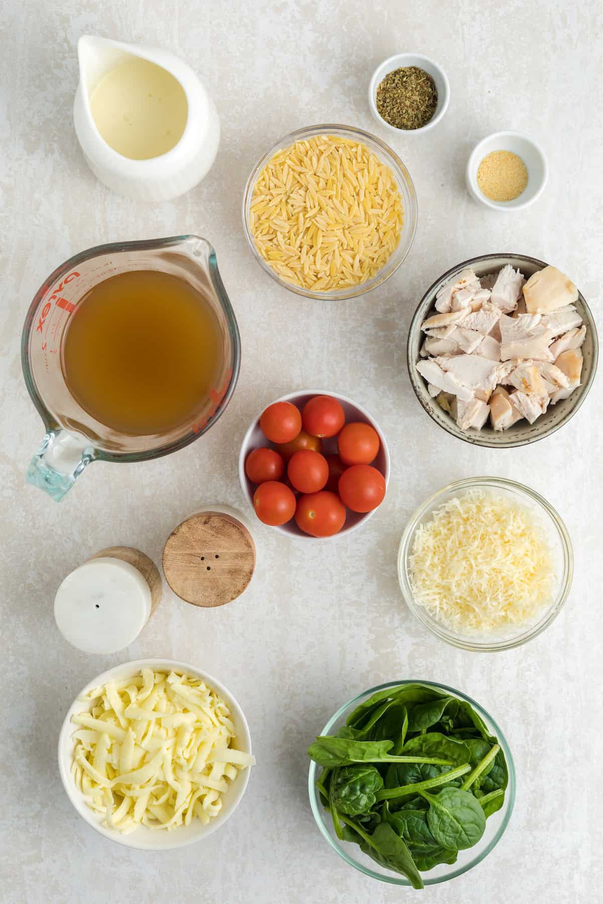 Ingredients for creamy chicken orzo pasta bake including spinach, cherry tomatoes, cooked chicken, shredded cheese, orzo, broth, and cream arranged on a light surface.