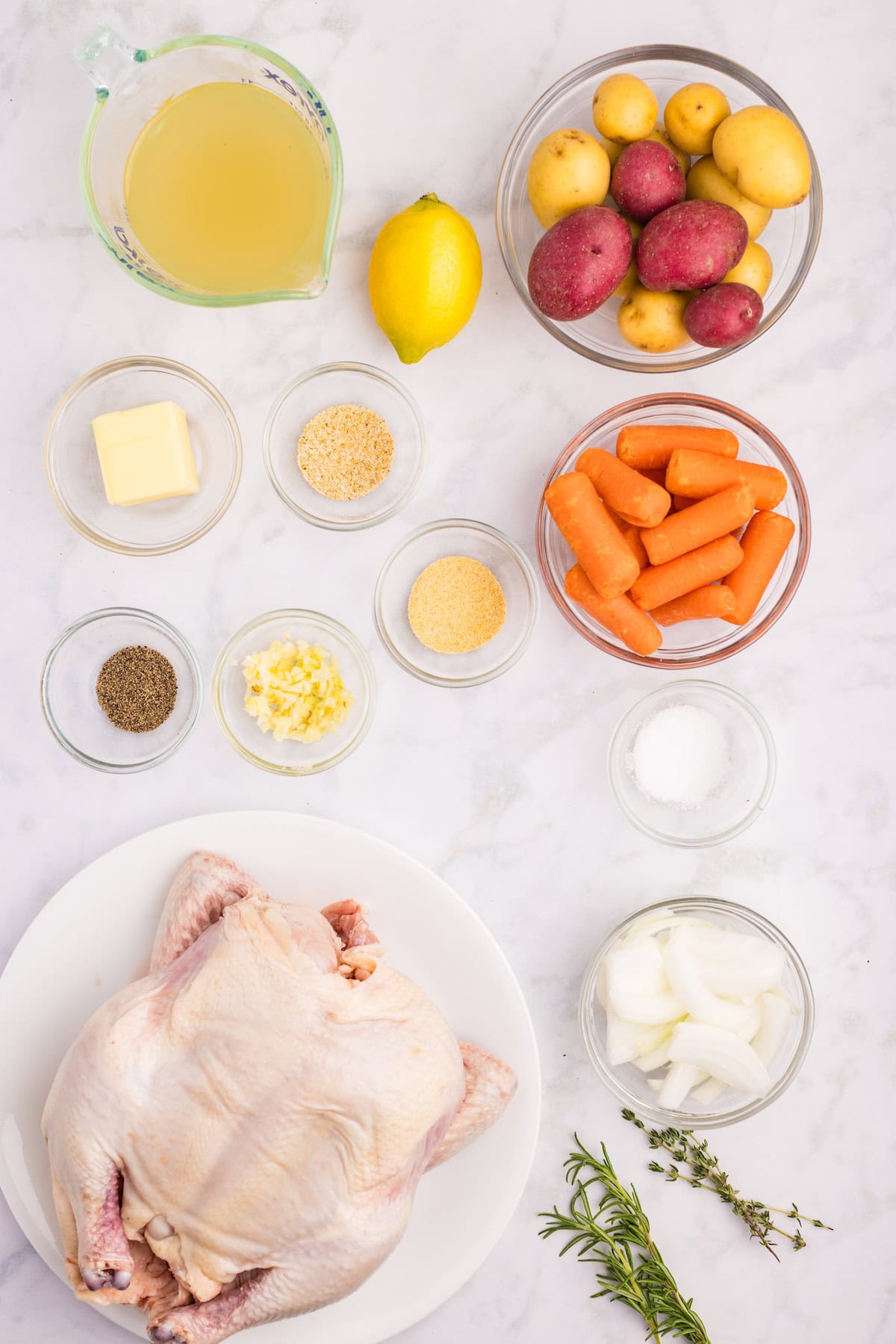 Overhead view of ingredients for herb roasted chicken, including a whole raw chicken, baby potatoes, carrots, lemon, butter, garlic, onion, herbs, and chicken broth arranged on a marble surface.