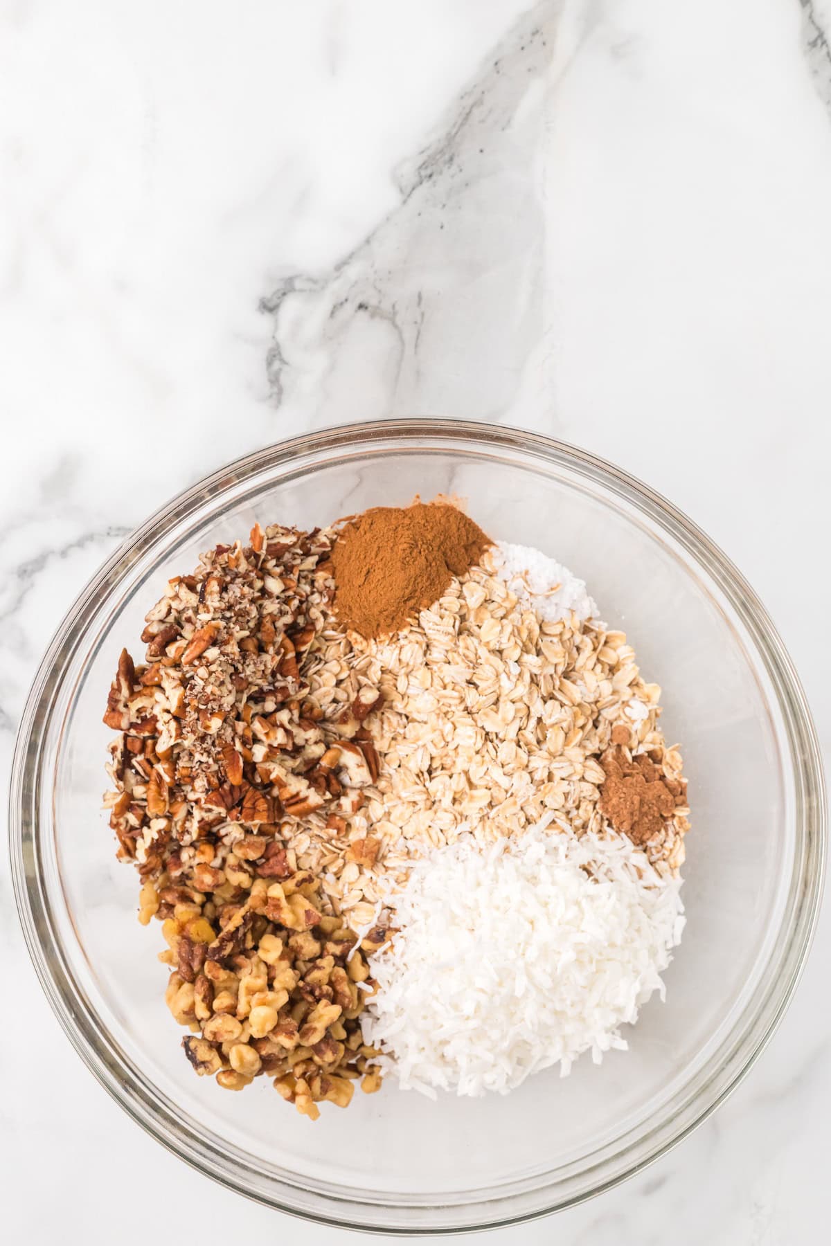 Glass bowl filled with rolled oats, chopped nuts, coconut flakes, cinnamon, and nutmeg before mixing to make homemade apple pie granola.