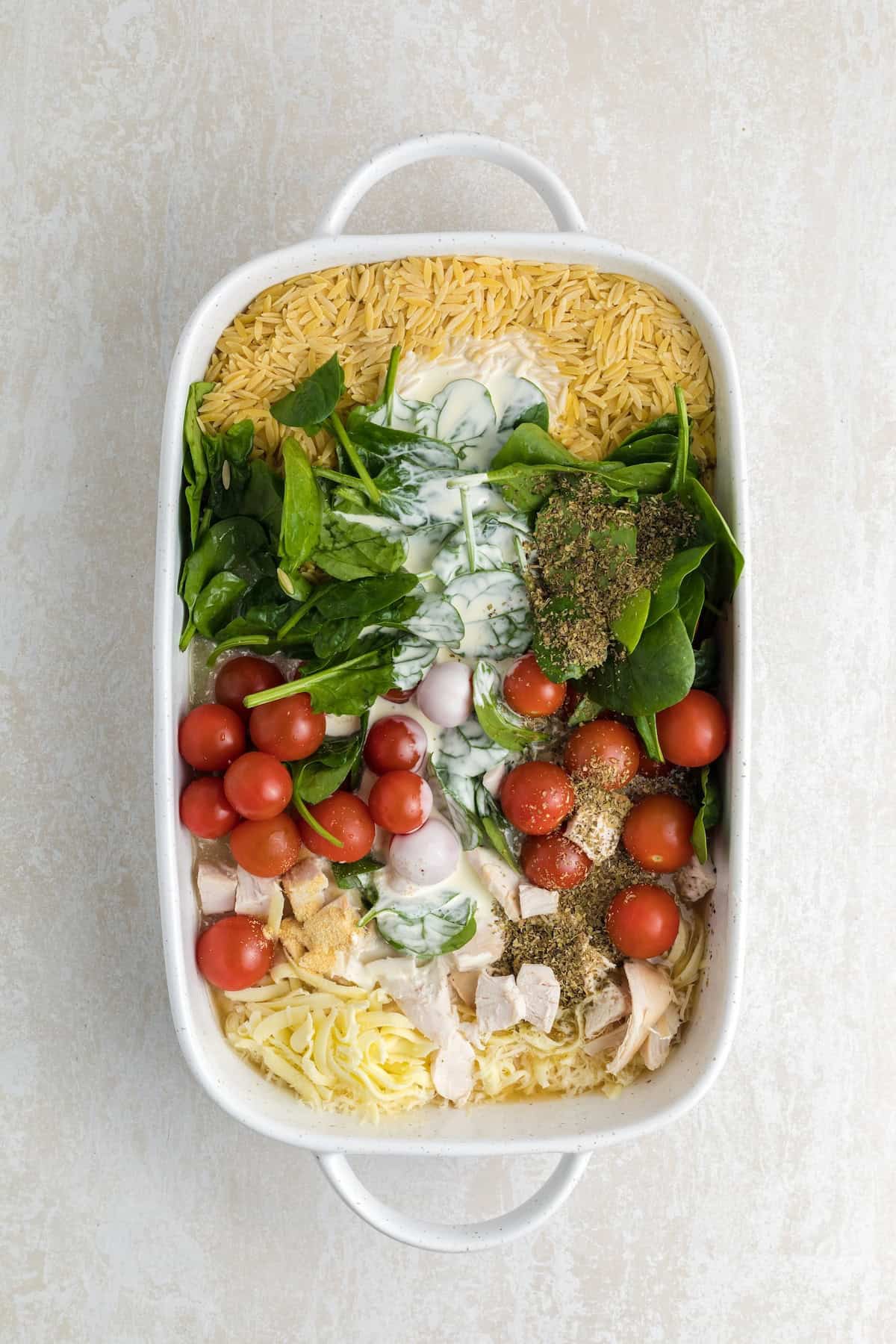 One-pan creamy chicken orzo mixture with spinach and cherry tomatoes combined in a white baking dish, ready to bake.