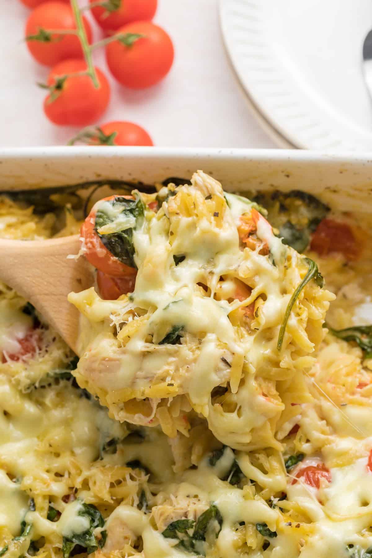 Close-up of creamy baked chicken orzo being scooped from a casserole dish with melted cheese, spinach, and cherry tomatoes, showing the gooey, cheesy texture.