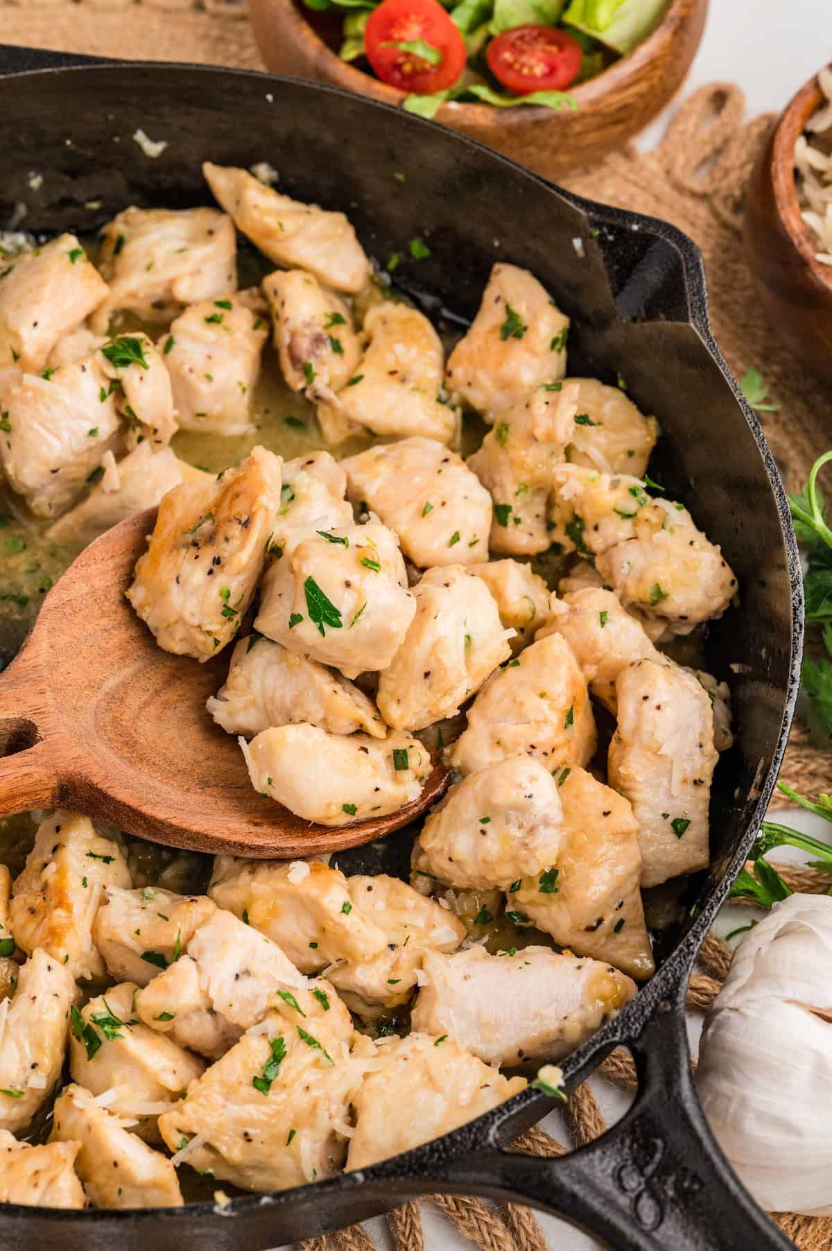 Close-up of One-Pan Garlic Parmesan Chicken Bites in a cast iron skillet. A wooden spoon lifts tender, golden chicken pieces coated in a buttery garlic parmesan sauce and sprinkled with fresh parsley, with a small salad bowl in the background.