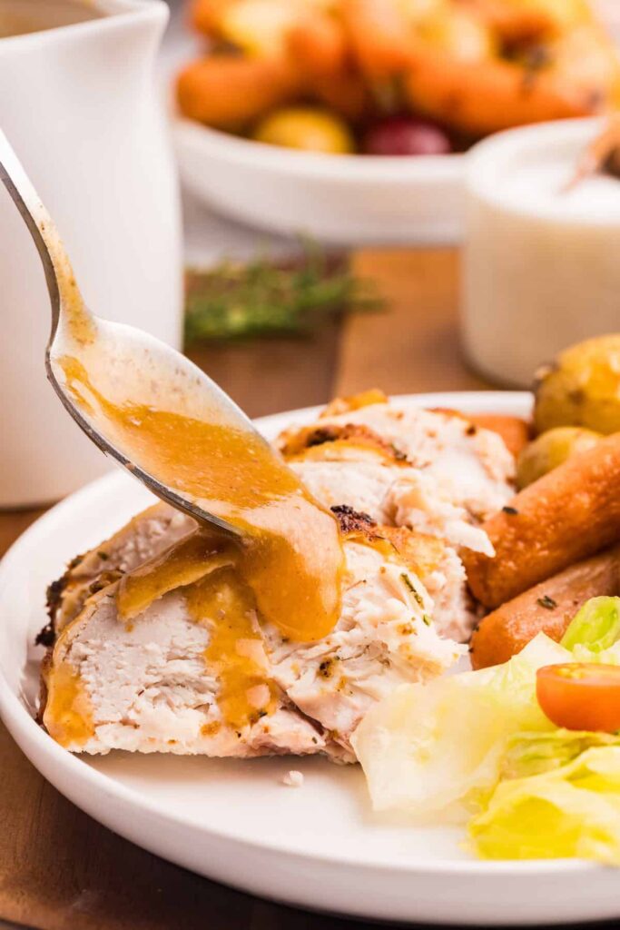 Close-up of golden gravy being spooned over sliced herb roasted chicken on a white plate with carrots, potatoes, and a fresh green salad.