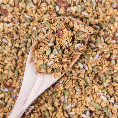Close-up of homemade pumpkin spice granola on a wooden spoon, showing clusters of oats, pumpkin seeds, sunflower seeds, and pecans with a golden, toasty coating.