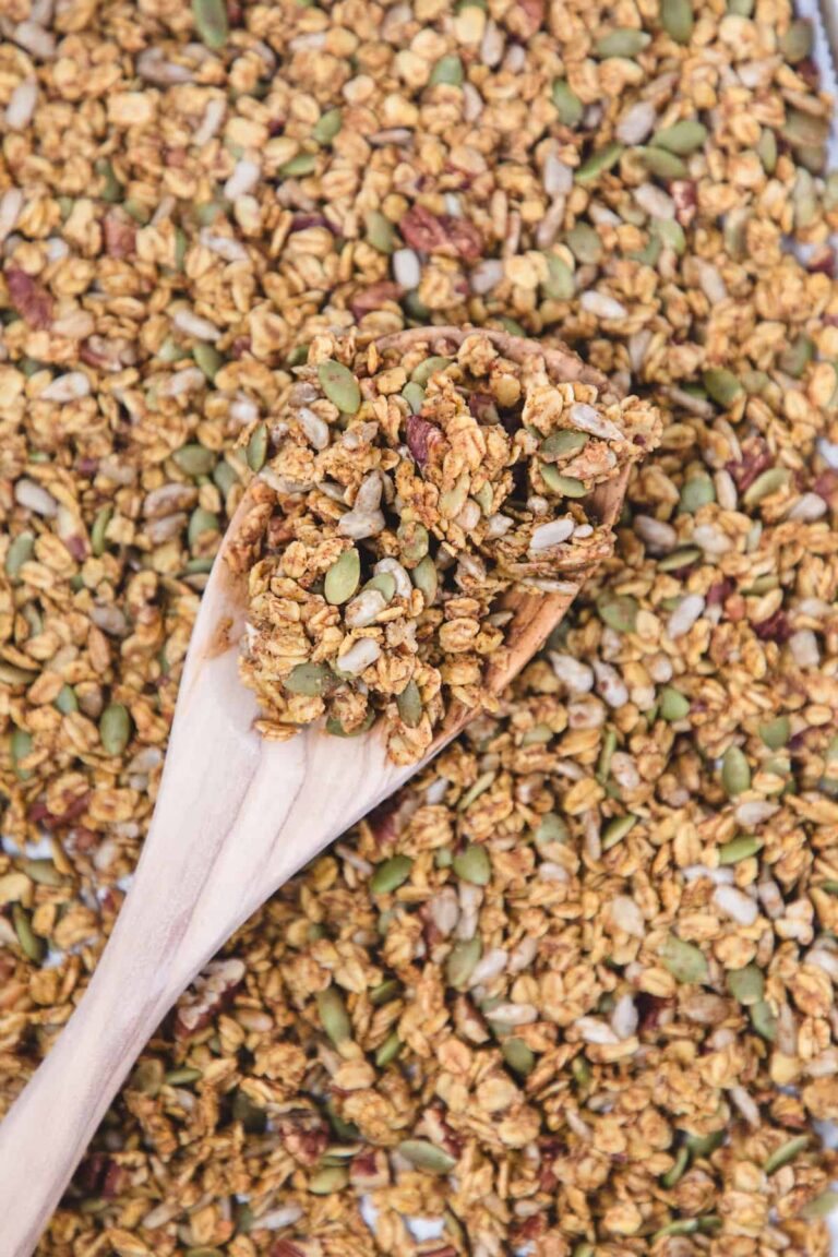 Close-up of homemade pumpkin spice granola on a wooden spoon, showing clusters of oats, pumpkin seeds, sunflower seeds, and pecans with a golden, toasty coating.