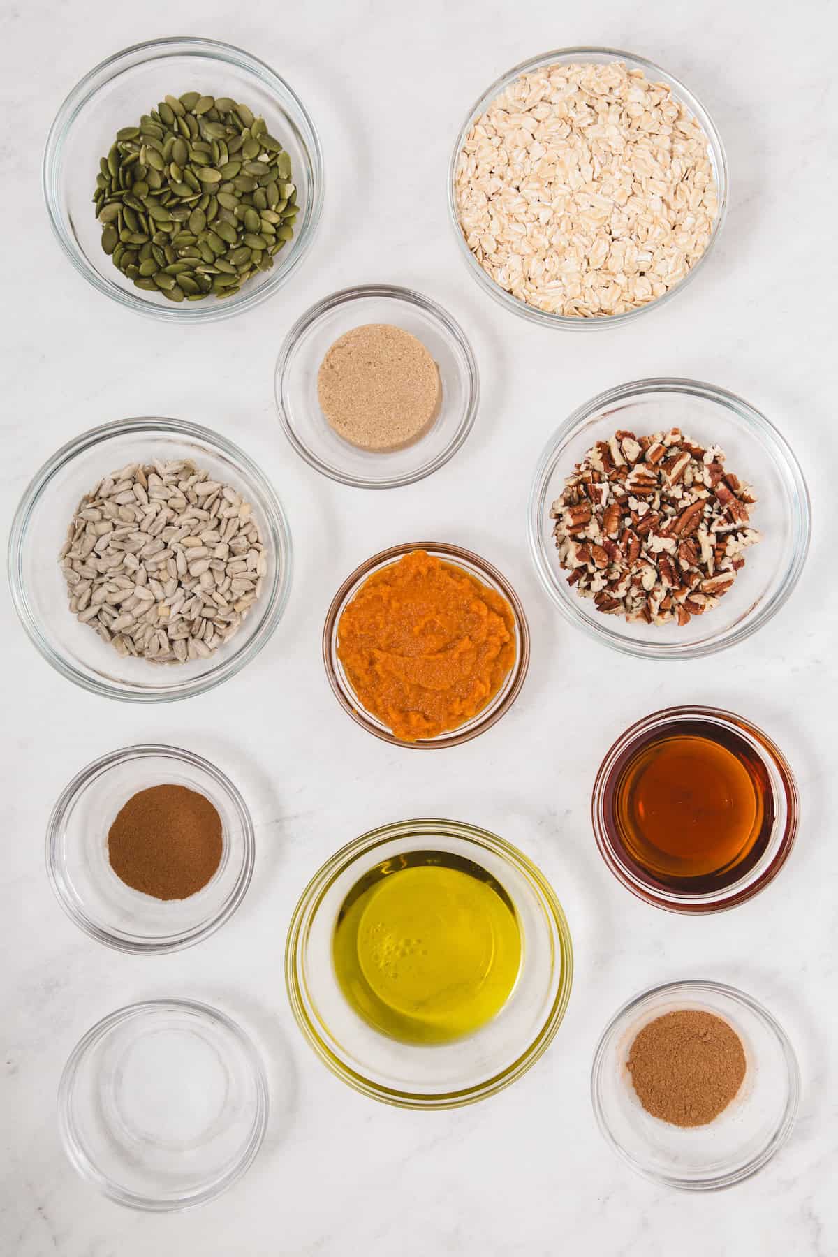 Overhead view of glass bowls filled with ingredients for pumpkin spice granola, including rolled oats, pumpkin seeds, sunflower seeds, chopped pecans, pumpkin puree, maple syrup, olive oil, brown sugar, pumpkin pie spice, cinnamon, and salt, all arranged neatly on a white surface.