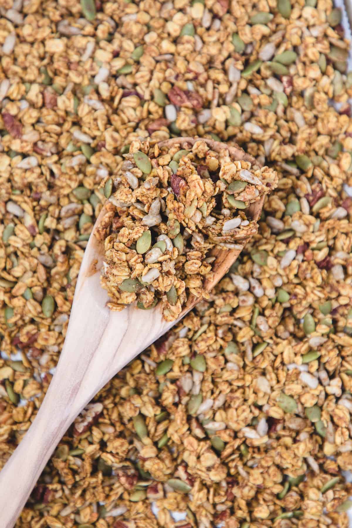 Close-up of homemade pumpkin spice granola on a wooden spoon, showing clusters of oats, pumpkin seeds, sunflower seeds, and pecans with a golden, toasty coating.