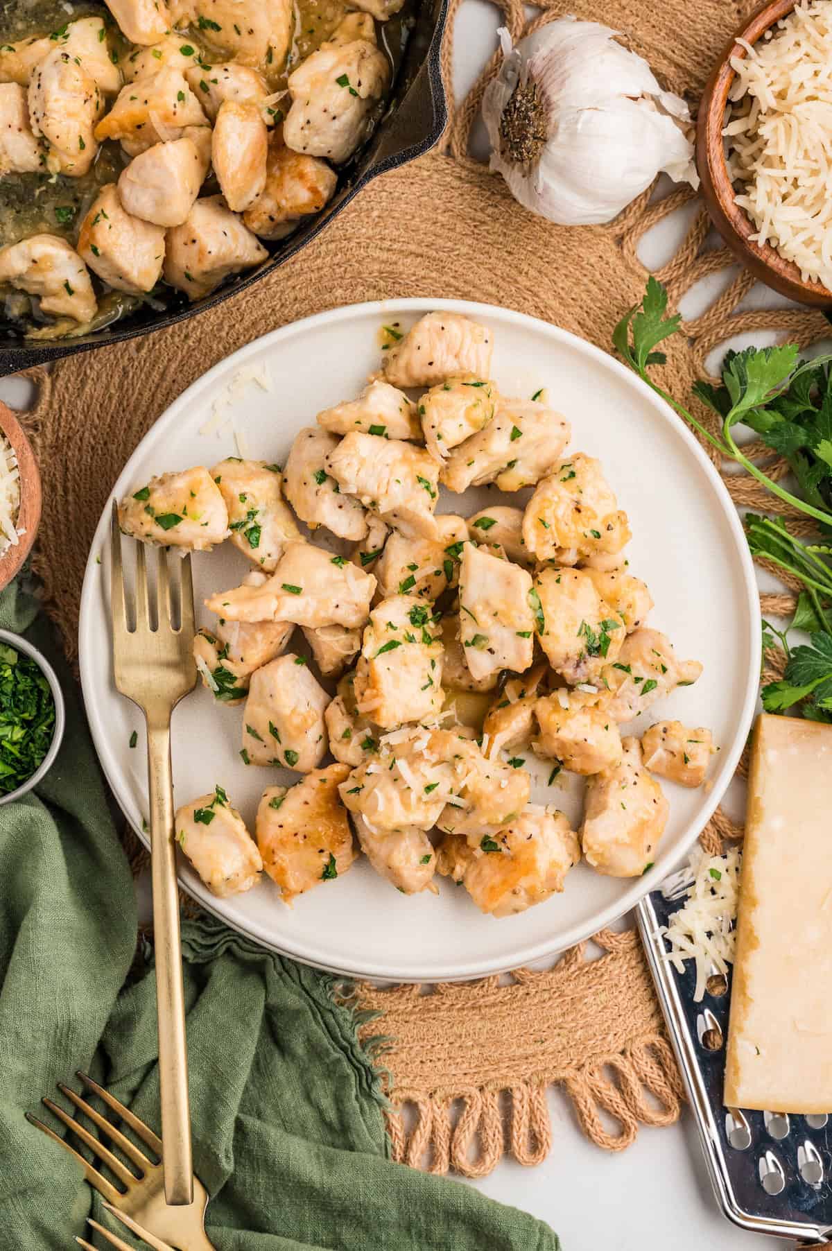Top-down view of Garlic Parmesan Chicken Bites served on a white plate with a gold fork. The golden, buttery chicken pieces are topped with chopped parsley and shredded parmesan. Surrounding the plate are fresh garlic, grated cheese, parsley, and a cast iron skillet for a cozy, rustic presentation.