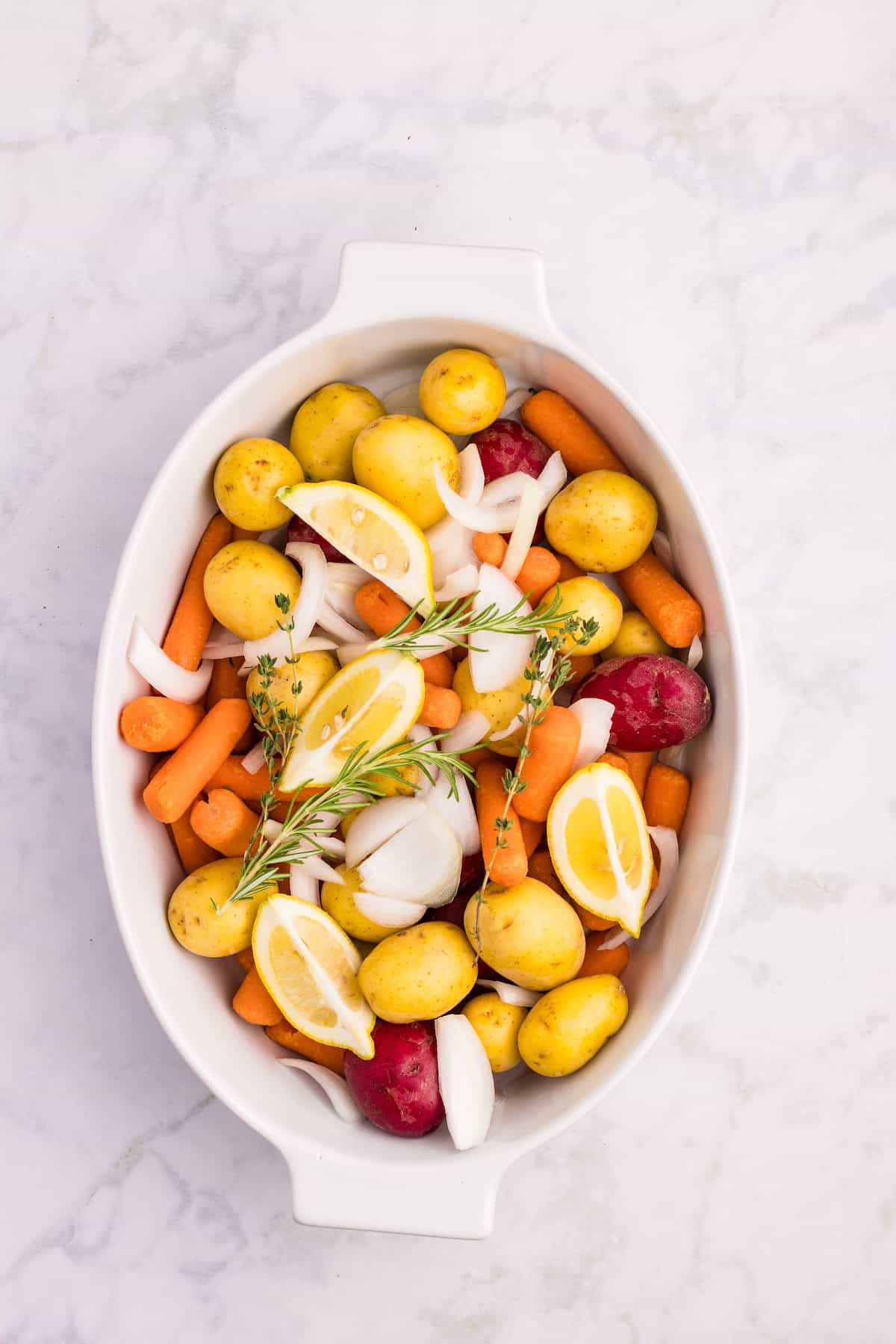 Baby potatoes, carrots, onion slices, lemon wedges, and fresh herbs arranged in a white baking dish, ready for roasting with herb butter chicken.