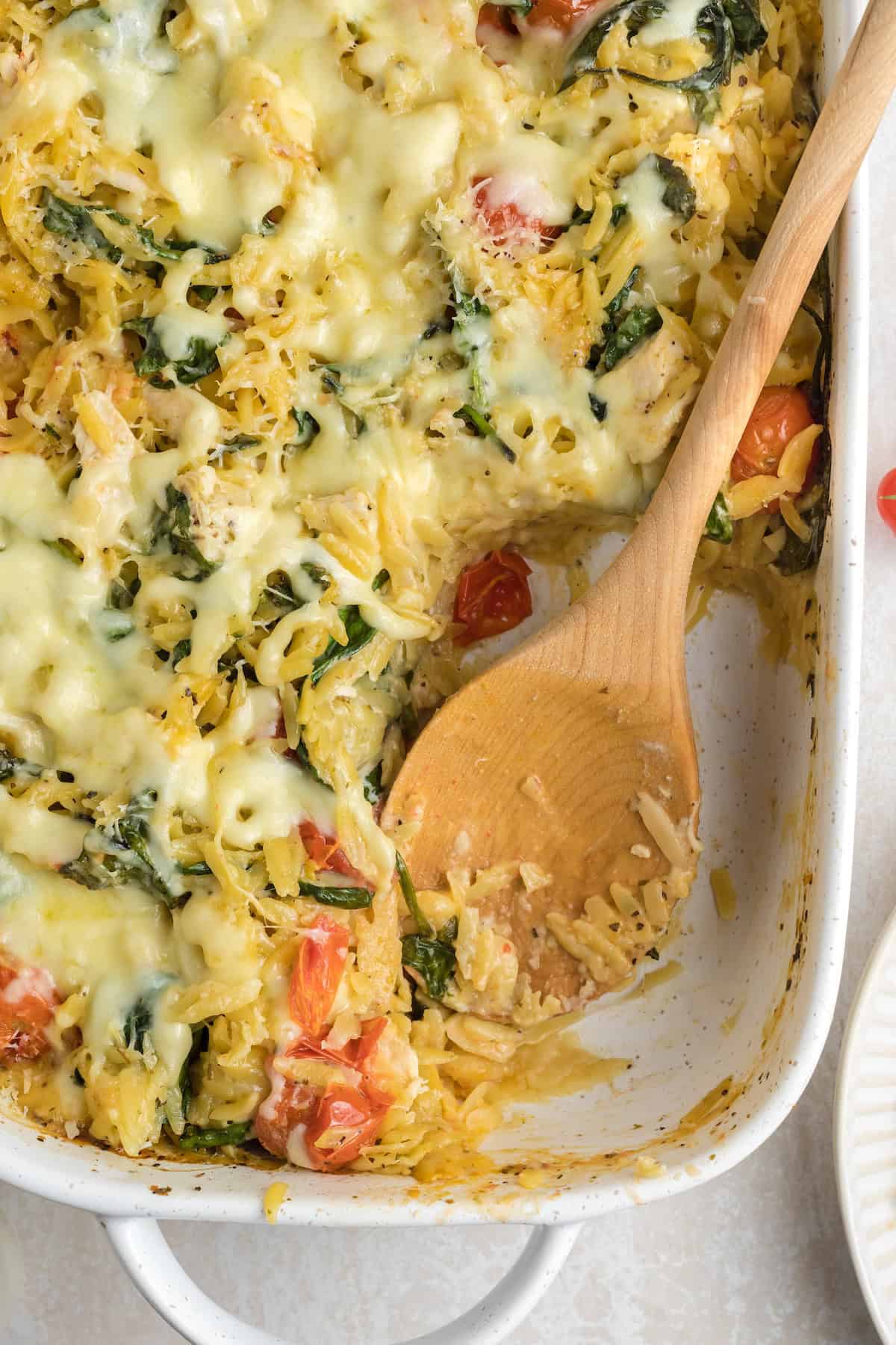 Serving creamy chicken orzo pasta bake with melted cheese, spinach, and tomatoes using a wooden spoon.