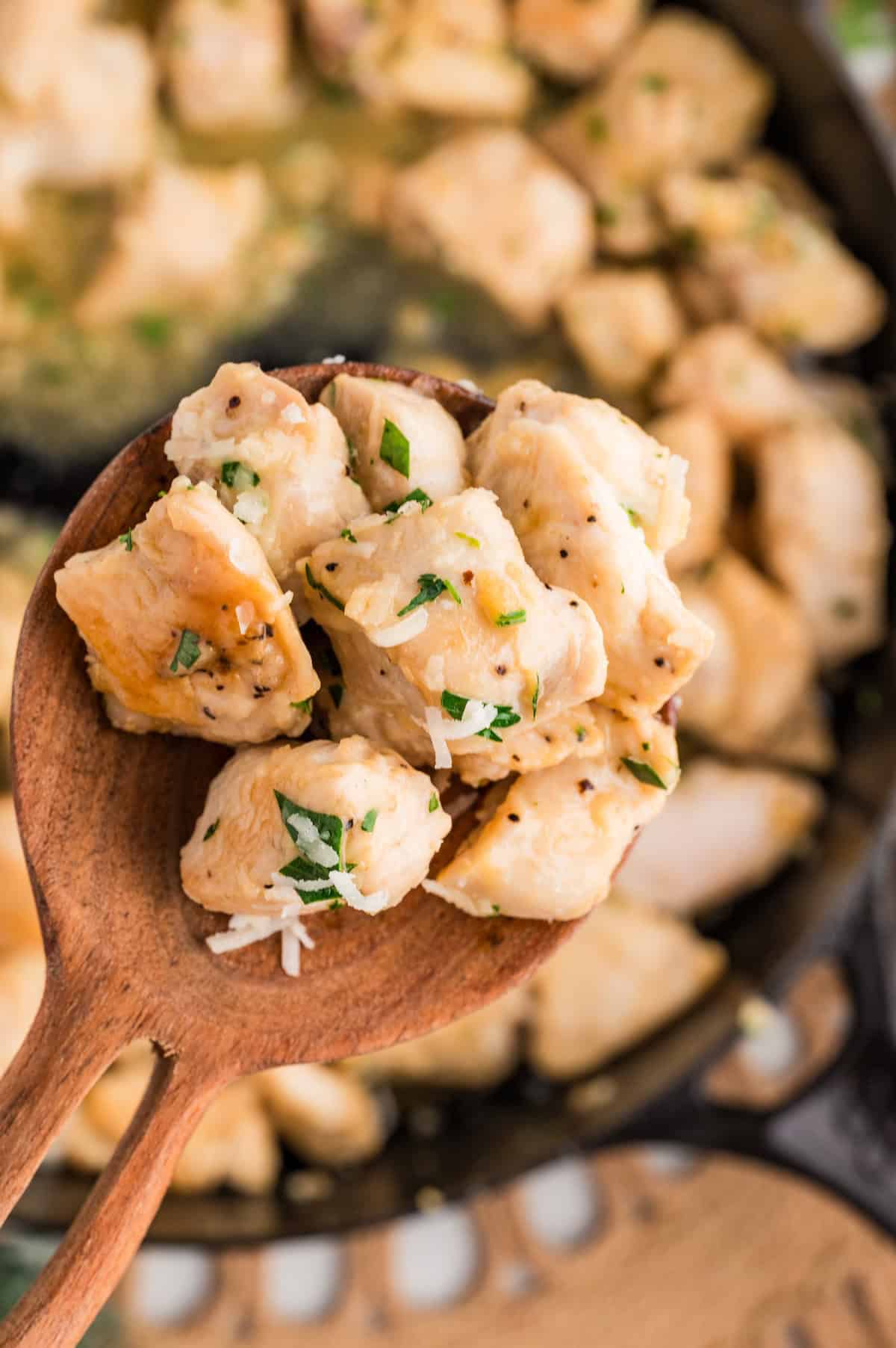 Close-up of a wooden spoon lifting tender Garlic Parmesan Chicken Bites from a skillet. The golden chicken pieces are coated in a buttery garlic parmesan sauce and sprinkled with chopped parsley and shredded parmesan cheese, highlighting their juicy texture and flavor.