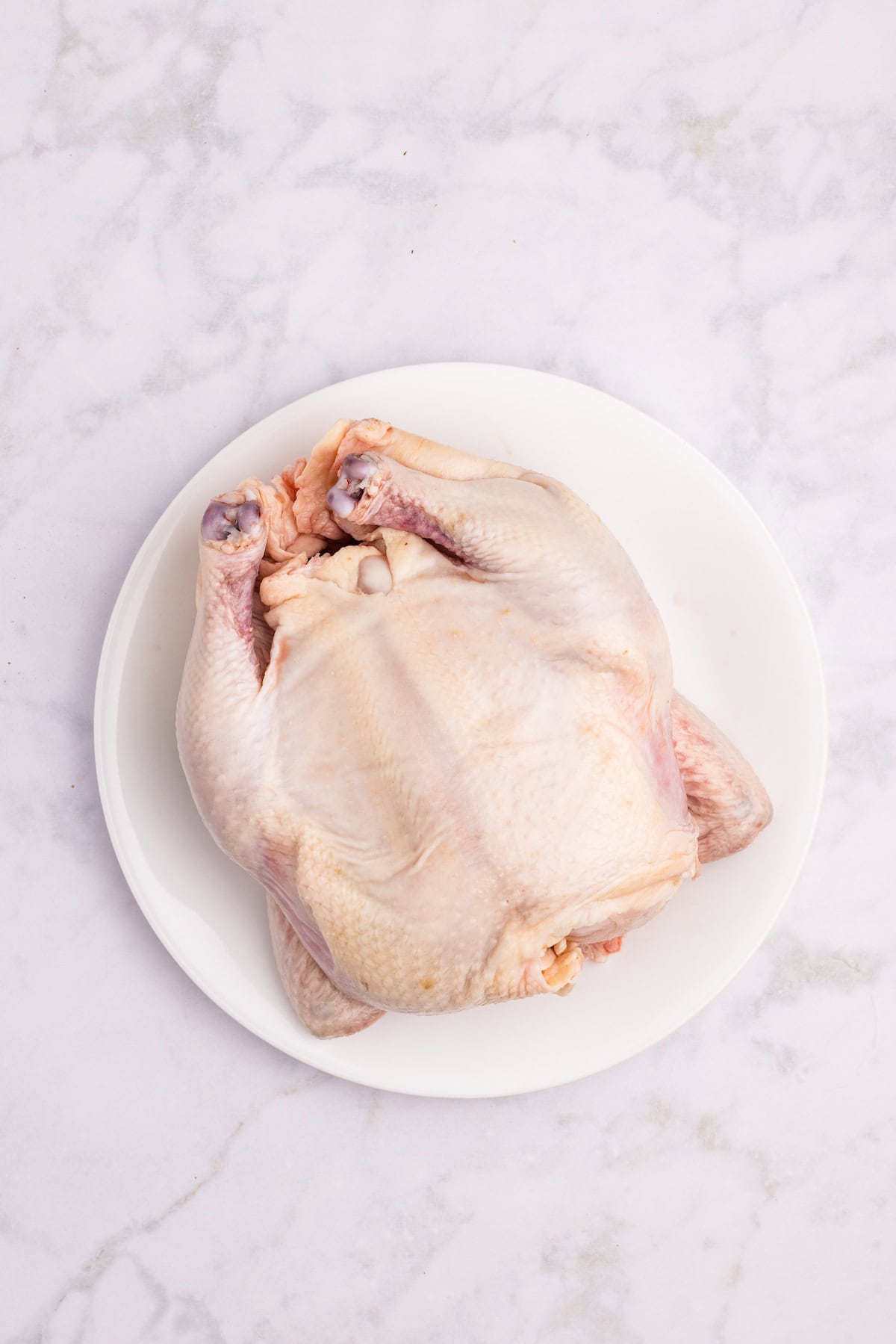 Unseasoned whole raw chicken on a white plate, prepped and ready to be rubbed with herb butter for roasting.