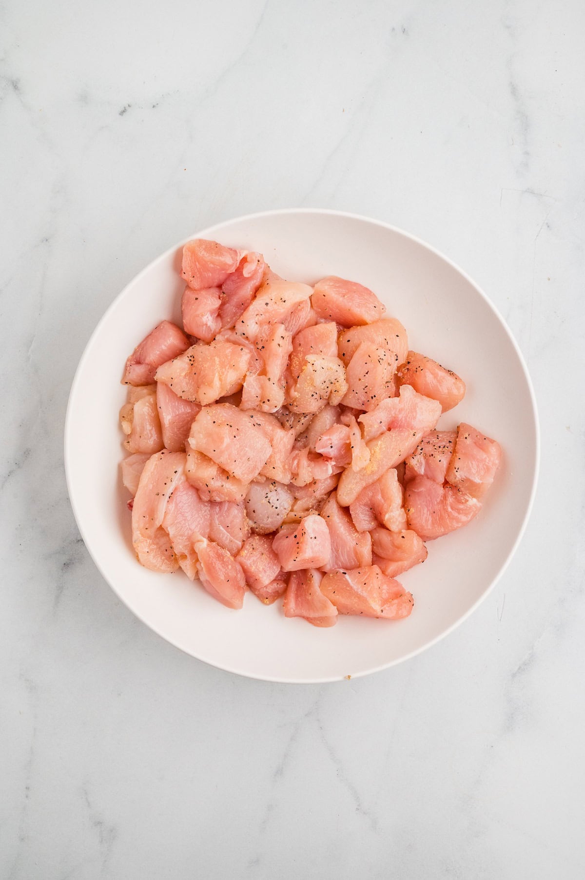 Top-down view of raw chicken breast pieces seasoned with salt, black pepper, and garlic powder in a white bowl on a marble surface, ready to be cooked for One-Pan Garlic Parmesan Chicken Bites.