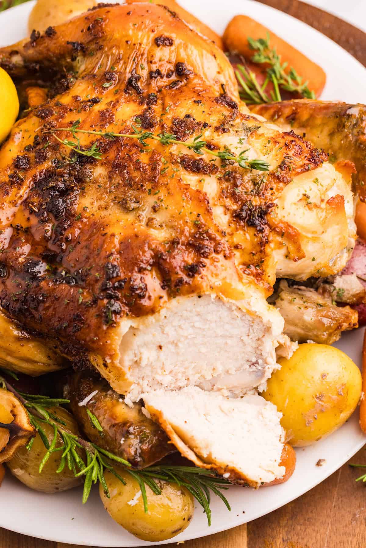 Close-up of juicy sliced herb roasted chicken surrounded by golden potatoes, carrots, and fresh rosemary on a white serving platter.