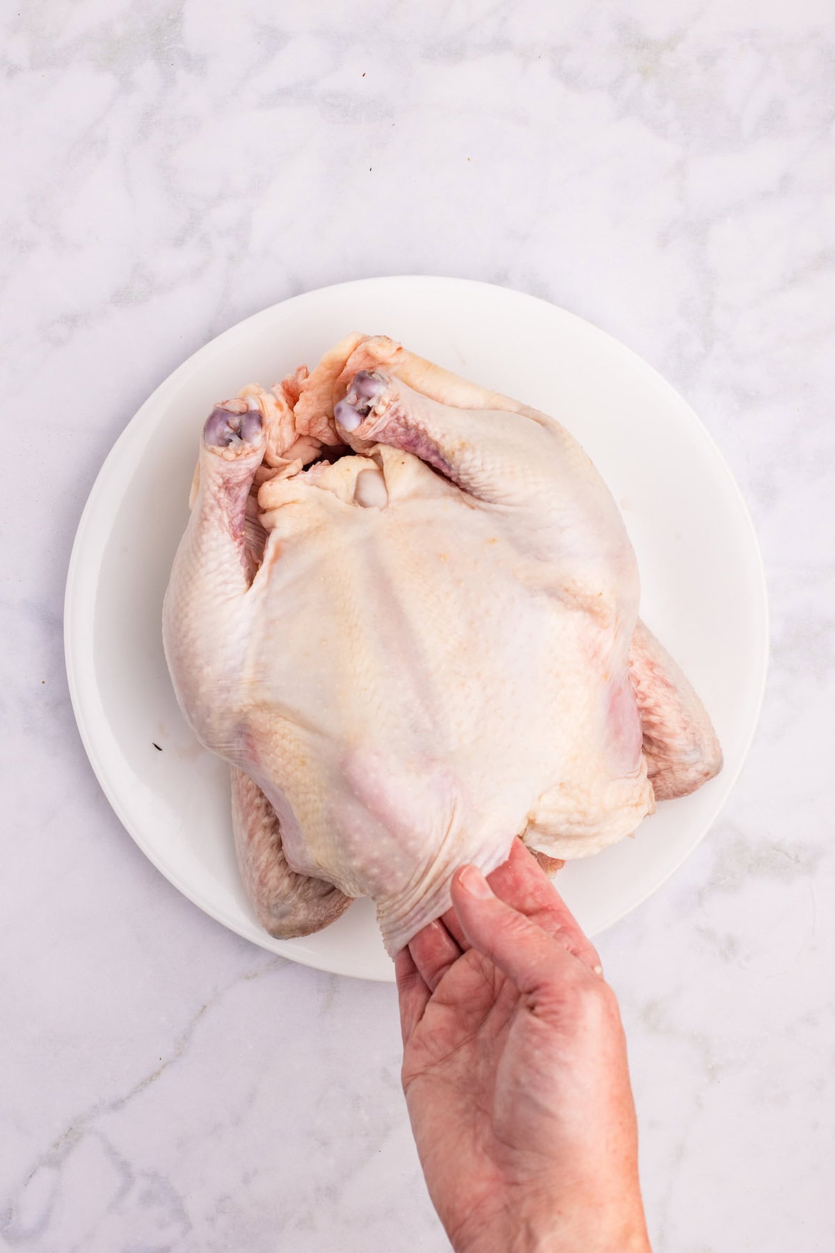 Hand gently lifting the skin of a whole raw chicken to spread butter underneath before roasting for tender, flavorful meat.