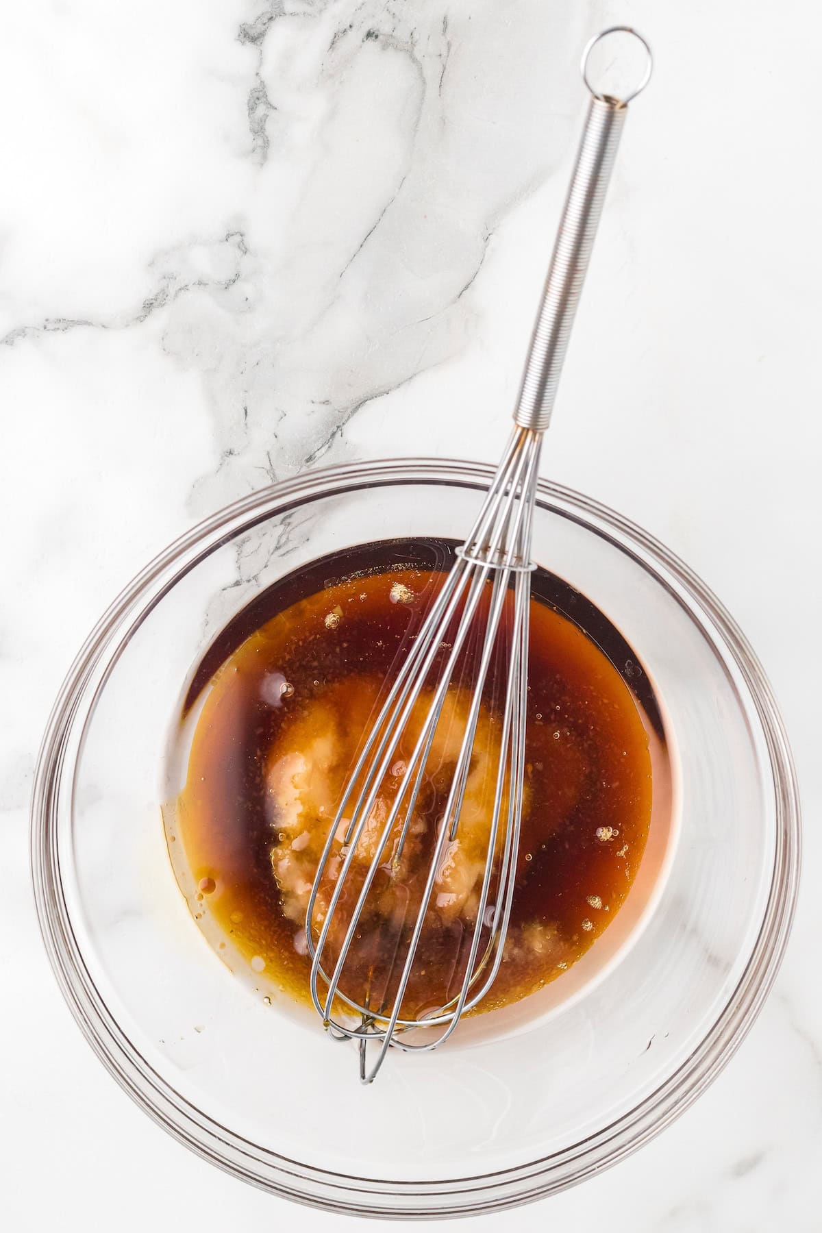 Glass bowl of maple syrup, applesauce, coconut oil, and vanilla being whisked together to create the wet mixture for apple pie granola.