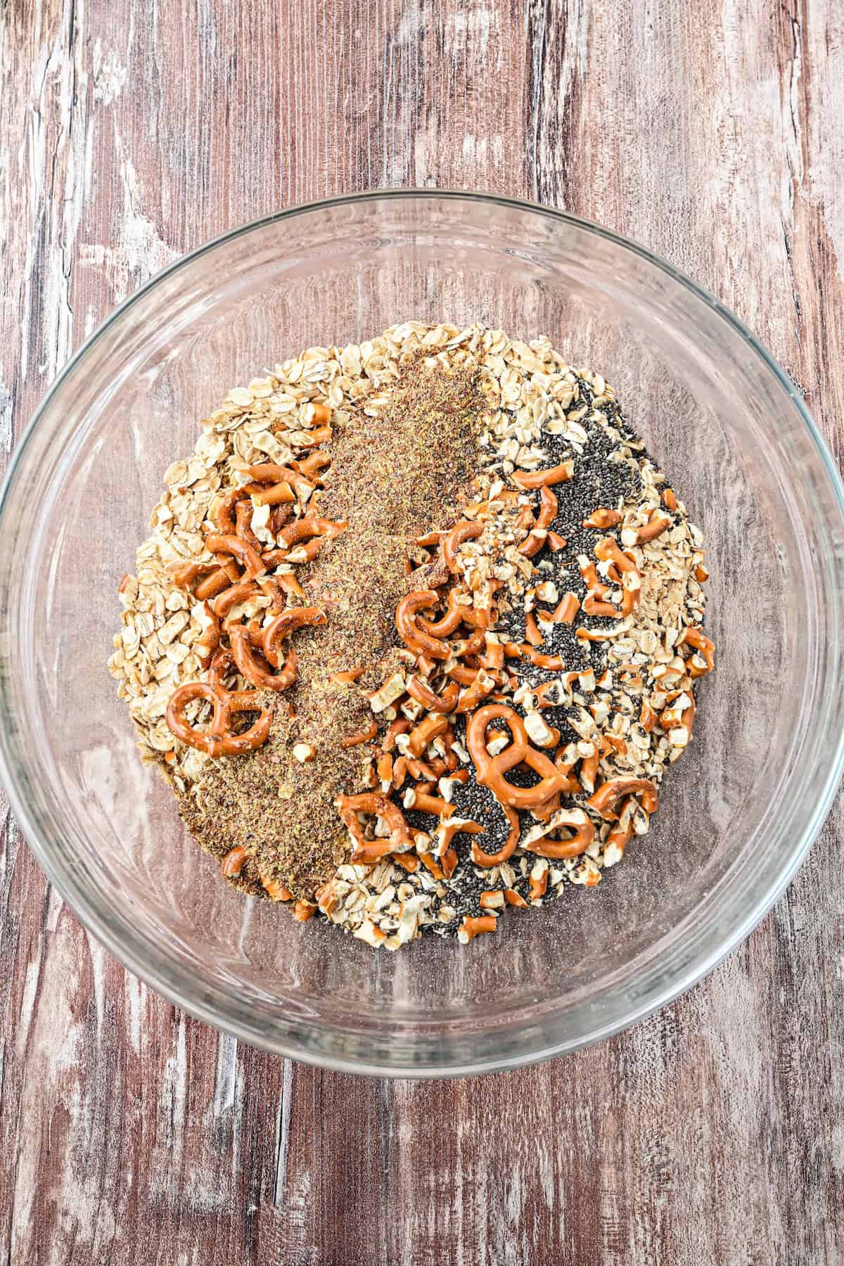 Overhead view of a glass bowl filled with dry ingredients for homemade chewy granola bars, including oats, crushed pretzels, chia seeds, and ground flaxseed.