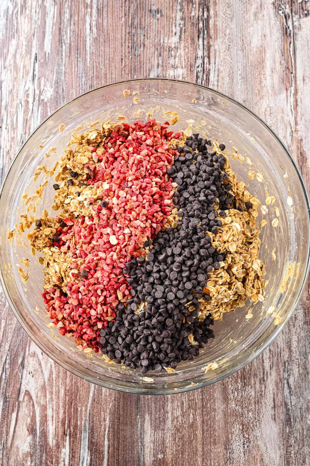 Overhead view of a glass bowl with granola bar mixture topped with freeze-dried strawberries and mini chocolate chips before mixing.