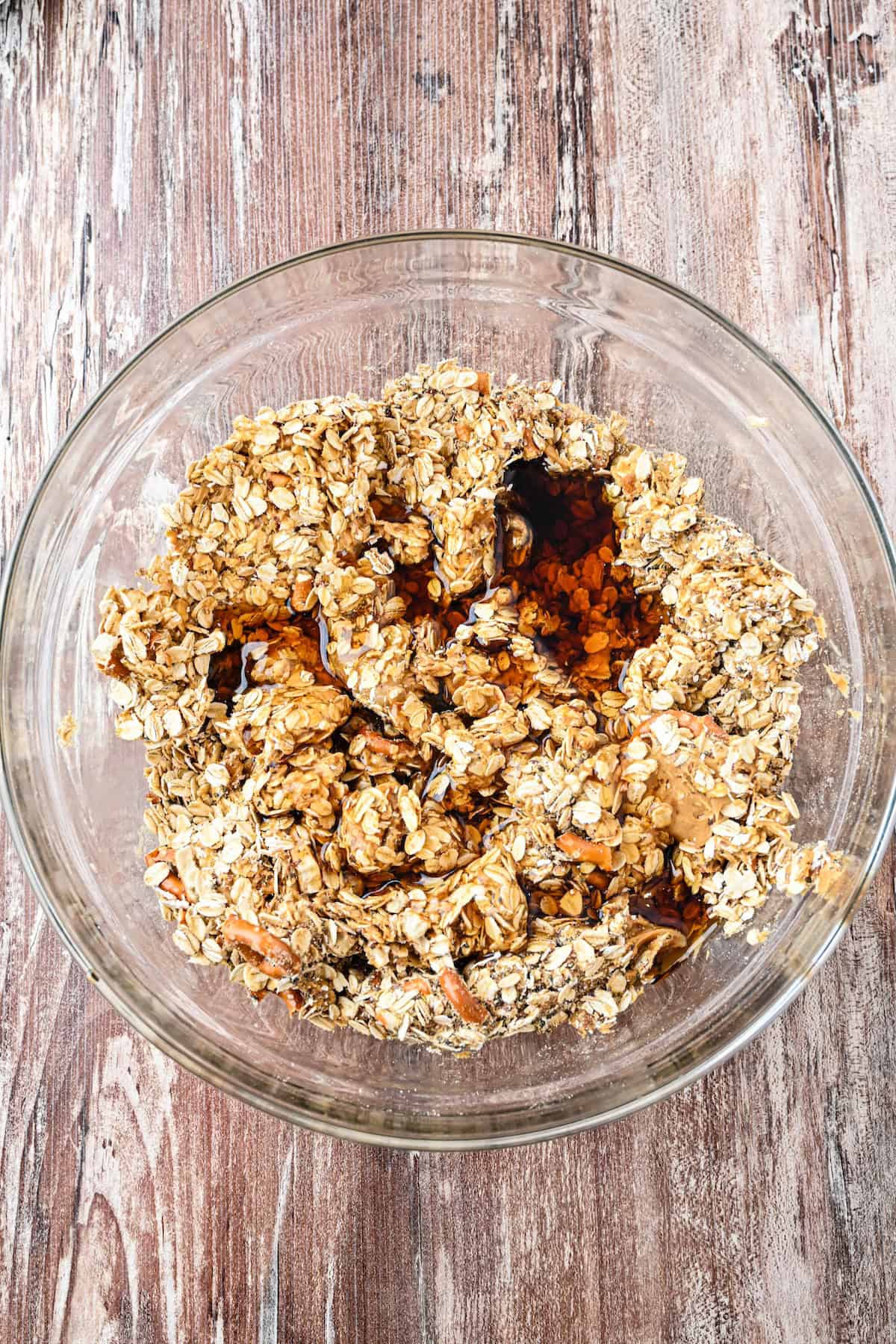 Overhead view of a glass bowl filled with oats and pretzels as maple syrup is added to the mixture for homemade no-bake granola bars.