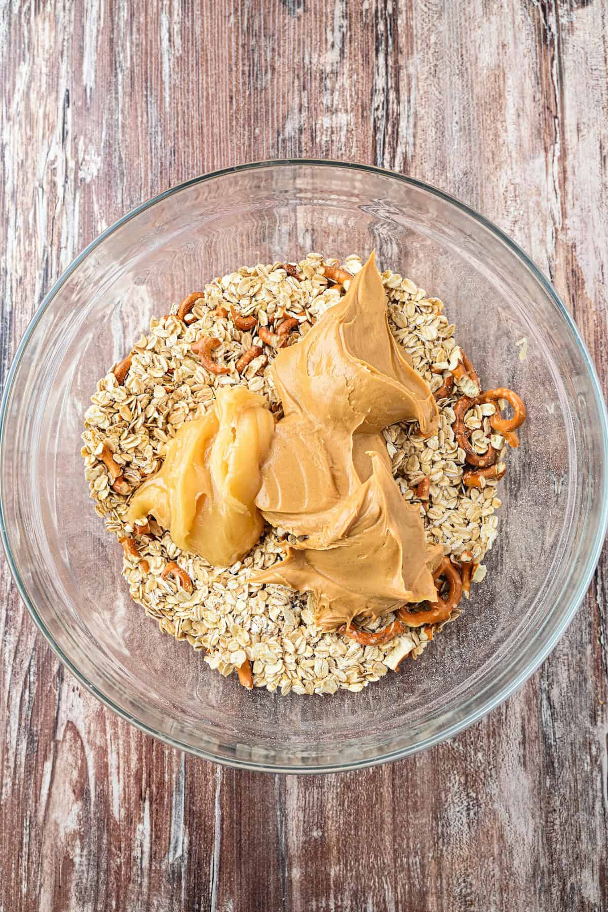 Overhead view of peanut butter and honey added on top of oats and pretzels in a glass bowl to make homemade chewy granola bars.