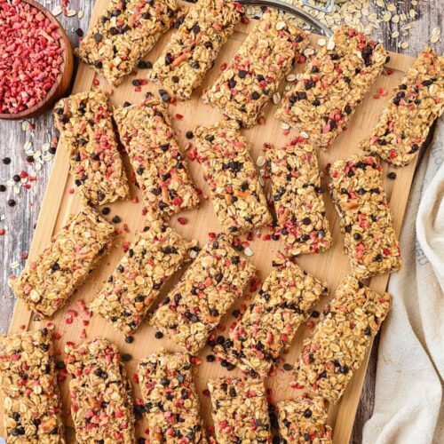 Overhead view of homemade chewy granola bars arranged on a wooden board, surrounded by oats, freeze-dried strawberries, and chocolate chips for a styled presentation.