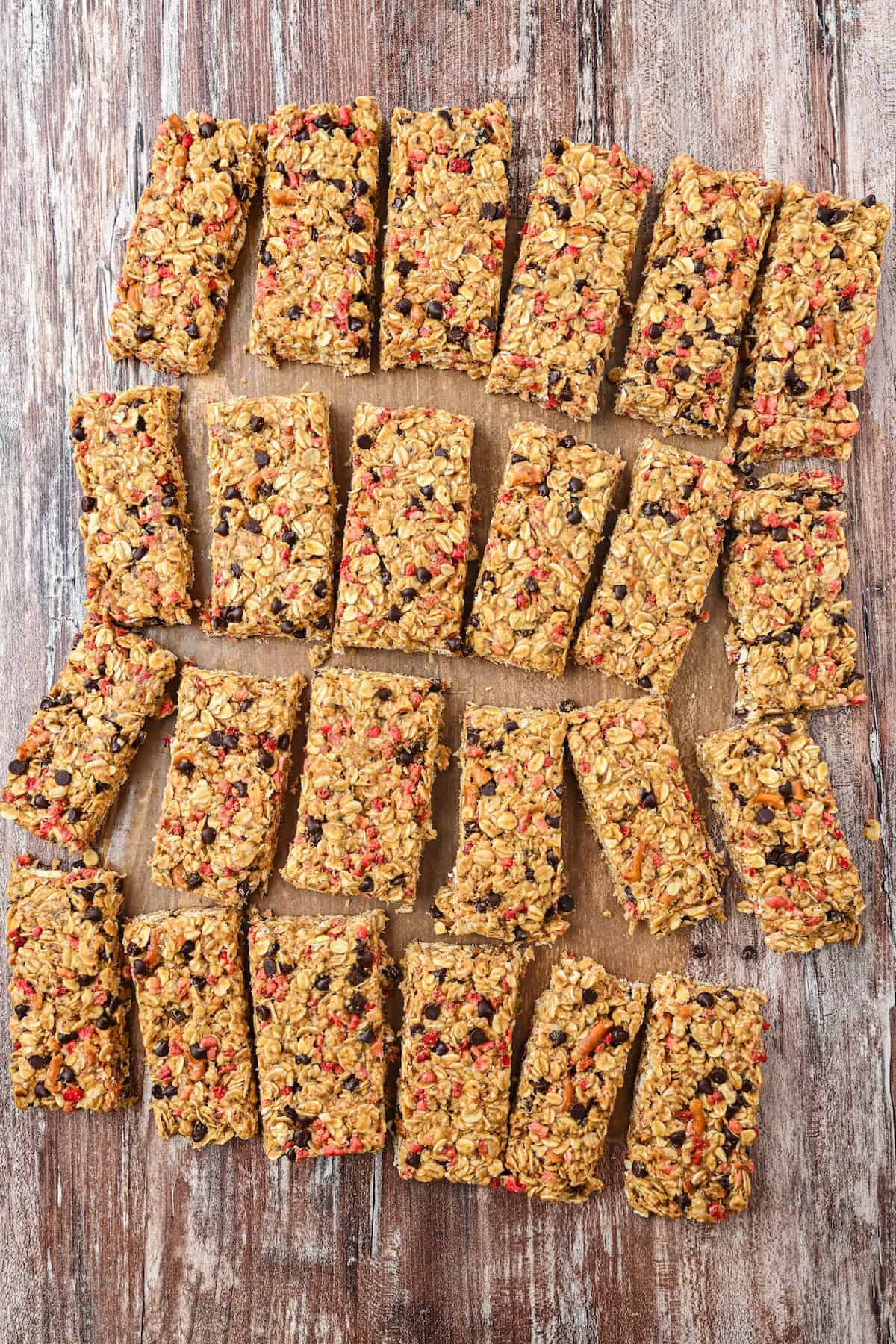 Overhead view of homemade chewy granola bars arranged on a wooden board, surrounded by oats, freeze-dried strawberries, and chocolate chips for a styled presentation.