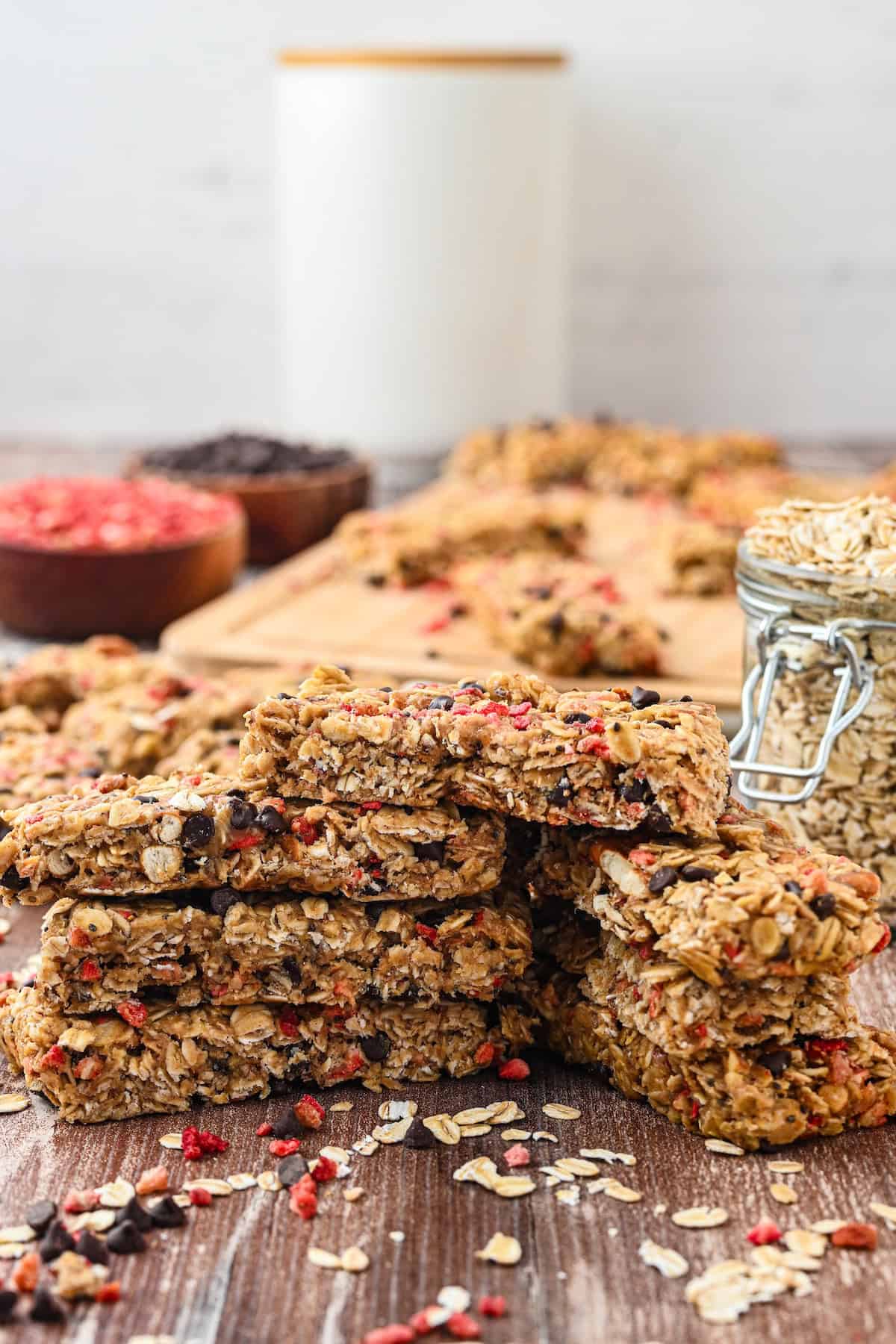Close-up view of a stack of homemade chewy granola bars filled with oats, chocolate chips, and freeze-dried strawberries, with additional bars and ingredients blurred in the background.