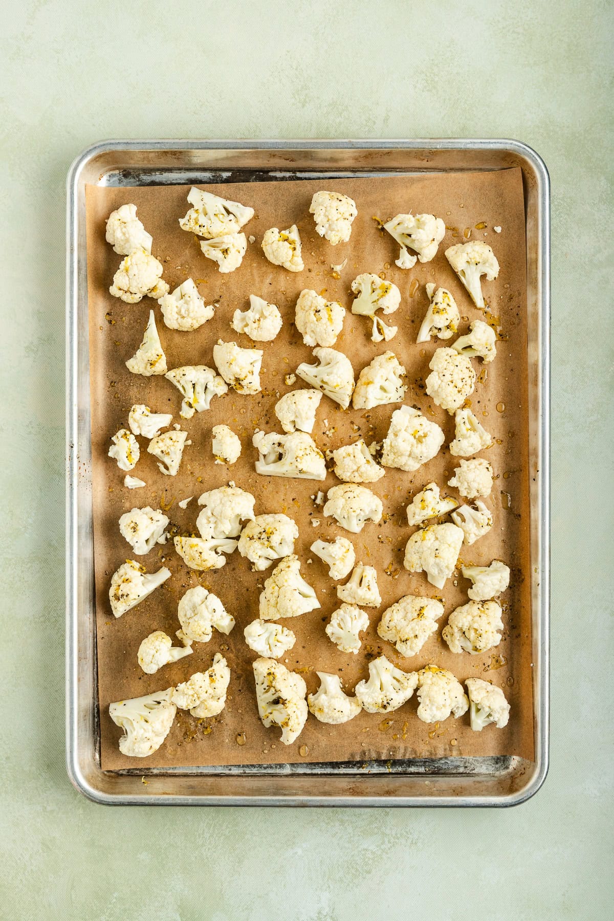 Raw cauliflower florets seasoned with olive oil, lemon zest, oregano, salt, and pepper on a parchment-lined baking sheet before roasting.