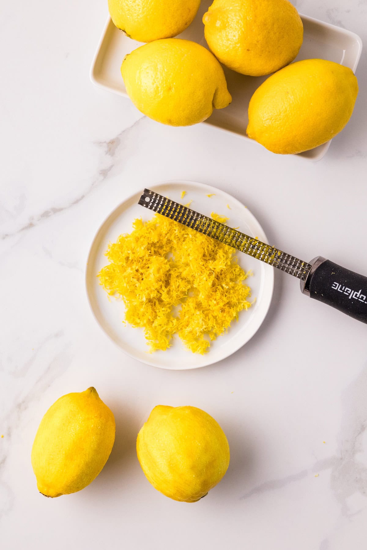 Fresh lemon zest on a plate with a microplane zester and whole lemons on a marble countertop, preparing ingredients for homemade lemon curd.